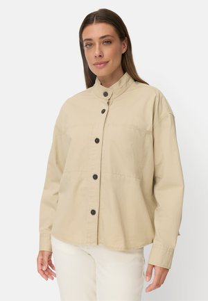 Vrouw met lang bruin haar, een beige jasje met knopen en opstaande kraag en een witte broek, staand tegen een effen achtergrond.