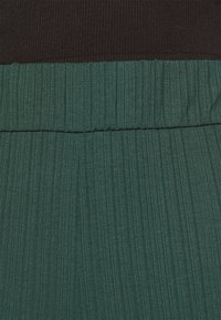 Lähedalt vaade tumerohelisele ribakanga küljele, millel on volditud vööosa, tumedast pruunist tekstuuriga kanga osal.