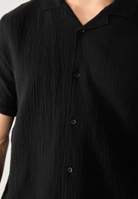 Camisa negra de manga corta, hecha de un tejido texturizado, con un acabado suave y arrugado, y un cuello con puntas decorado con seis botones.