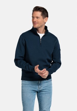 Dunkelblauer Half-Zip-Pullover aus weichem Stoff, mit hohem Kragen, langen Ärmeln und gerippten Bündchen, kombiniert mit hellblauen Jeans.