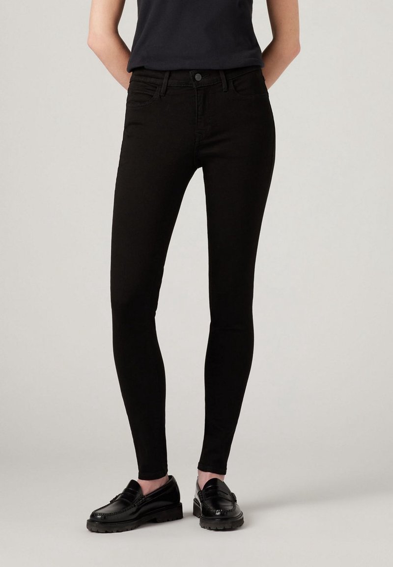 Personne portant un jean noir ajusté associé à des mocassins noirs, debout devant un fond uni gris clair.