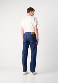 Vaqueros de mezclilla azul oscuro con pierna recta, que presentan un parche de cuero marrón claro en la parte trasera de la cintura y un diseño de cinco bolsillos.
