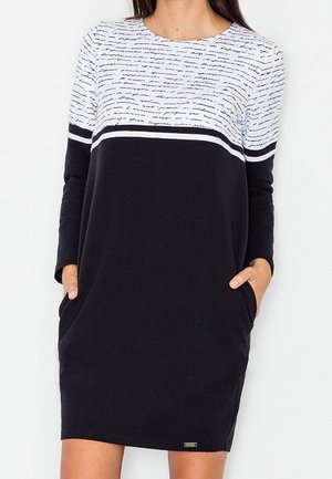 Woman wearing a long-sleeve black dress with a white top featuring cursive text and horizontal stripes, hands in side pockets.