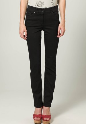 Frau trägt schwarze Slim-Fit-Jeans und rote Plateau-Sandalen, steht vor einem neutralen Hintergrund.