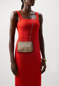 Metaliczna złota torebka crossbody o teksturowanej powierzchni, w połączeniu z dopasowaną czerwoną sukienką z grubymi ramiączkami i wyraźnie zaznaczoną talią.