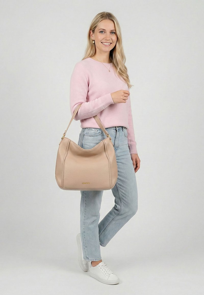 Mujer rubia sonriendo, vestida con un suéter rosa y vaqueros claros, sosteniendo un bolso beige sobre el hombro, de pie sobre un fondo claro.