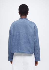 Oseba, pogledana od zadaj, nosi modro denim jakno z dolgimi rokavi, prirejeno do pasu, ter bele hlače na svetli enobarvni podlagi.