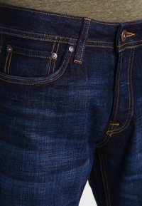 Dunkelblaue Jeans aus Denim mit orangefarbenen Nähten, ausgestattet mit vorderen Taschen und Metallbeschlägen. Der Stoff hat eine glatte Textur mit dezenten Farbnuancen.