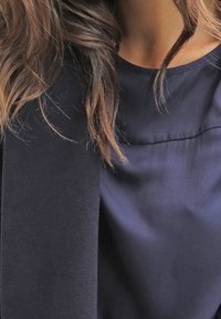Mørkeblå bluse med en glat, skinnende overflade, der har en rund halsudskæring og diskrete syning detaljer på brystet. Håret dækker delvist stoffet.