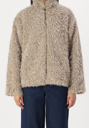 Beżowa miękka kurtka na zamek z wysokim kołnierzem, noszona z ciemnoniebieskimi, szerokimi dżinsami, widoczna od szyi do bioder na białym tle.