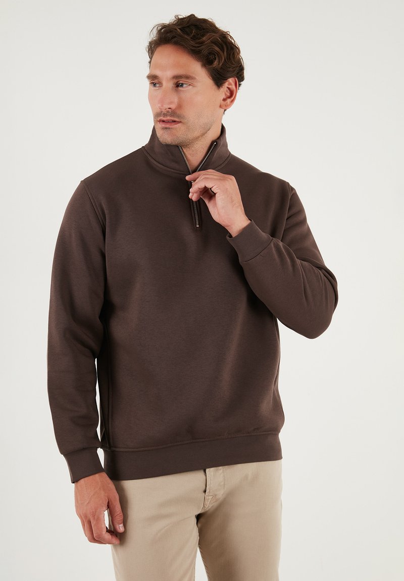 Bruine zip-necksweater met lange mouwen, zacht stof, ribgebreide manchetten en een naadloze afwerking. Gecombineerd met lichte broek.