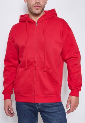 Homme portant un sweat à capuche rouge à fermeture éclair avec poches avant et cordons, associé à un jean bleu.