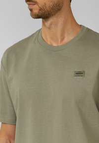 Olijfgroen katoenen T-shirt met korte mouwen, geribde ronde hals en een klein rechthoekig logoplaatje op het linkerborstgebied.
