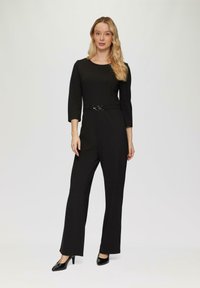 Zwarte jumpsuit met driekwart mouwen en een getailleerde taille, met een ronde metalen riem. Model draagt zwarte hoge hakken.