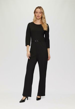 Zwarte jumpsuit met driekwart mouwen en een getailleerde taille, met een ronde metalen riem. Model draagt zwarte hoge hakken.