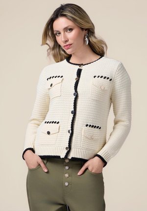 Cárdigan de textura cremosa con ribetes negros, que presenta dos bolsillos con botones y un cuello redondo. Combinado con pantalones verde oliva.