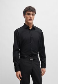 Maglietta nera a maniche lunghe con chiusura a bottoni, realizzata in tessuto liscio, abbinata a pantaloni scuri e una cintura nera con fibbia argentata.
