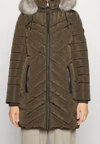 Olivgrün gesteppter Wintermantel mit Frontreißverschluss, handgefutterte Taschen mit schwarzem Rand, gerippte Bündchen und einer mit Fell verzierten Kapuze.