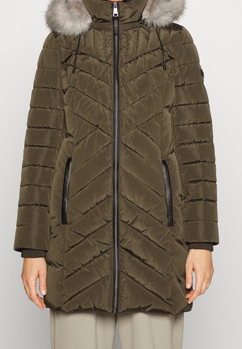 Olivgrün gesteppter Wintermantel mit Frontreißverschluss, handgefutterte Taschen mit schwarzem Rand, gerippte Bündchen und einer mit Fell verzierten Kapuze.