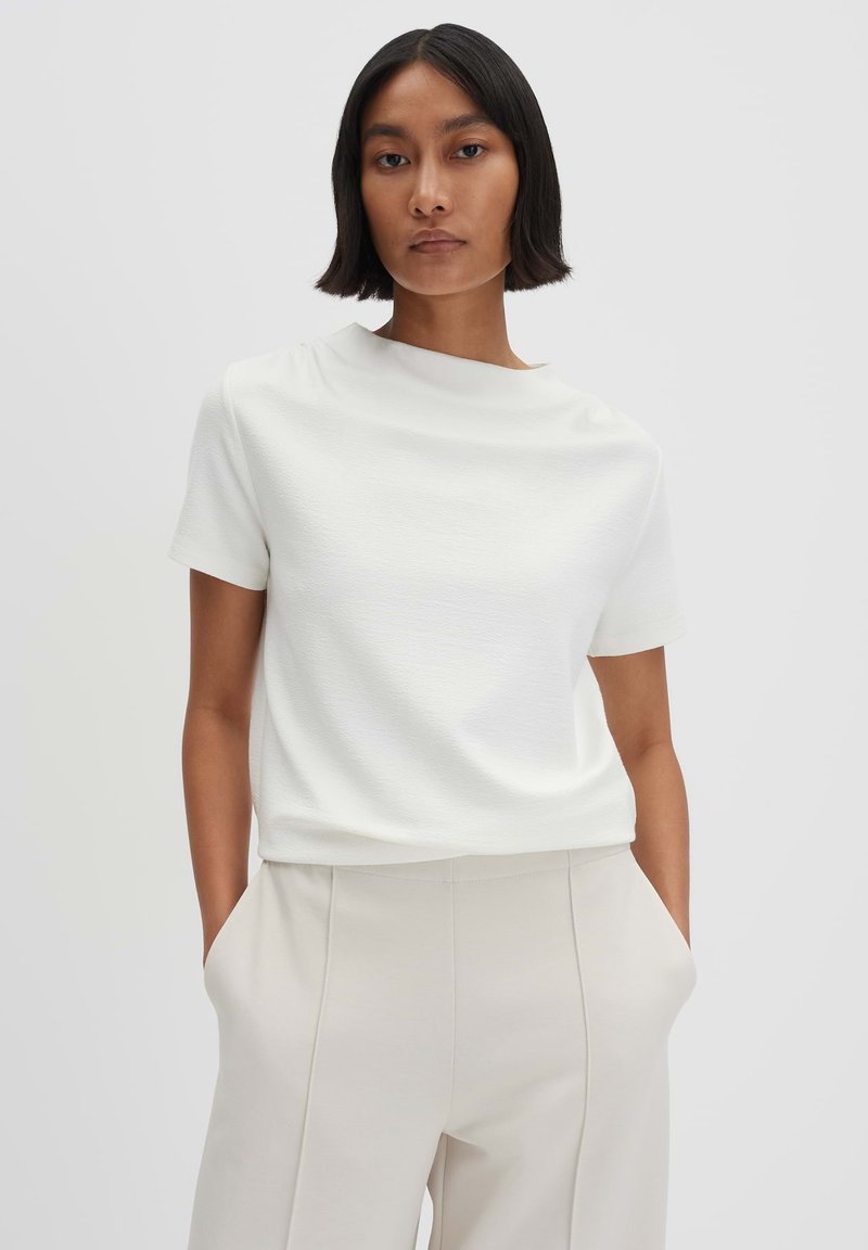Haut blanc à manches courtes avec une finition texturée et un col rond, associé à un pantalon taille haute beige clair avec des poches sur les côtés.