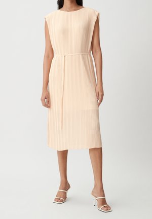 Kvinde iført en ærmeløs beige plisseret midi kjole med et tyndt bånd i taljen og hvide højhælede sandaler mod en ensfarvet baggrund.