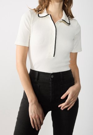 Hvid ribstrikket poloshirt med korte ærmer, sort kant og knapper, kombineret med højtaljede sorte jeans. Designet har krave og struktureret stof.