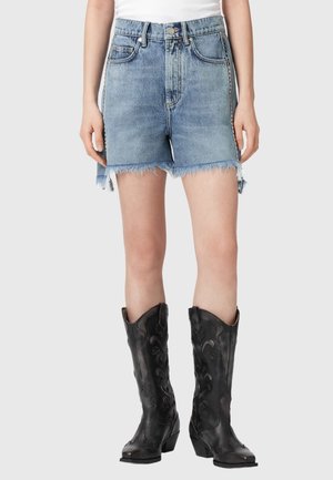 Femeie purtând pantaloni scurți din denim cu tiv destrămat și cizme de cowboy negre cu model, stând pe un fundal simplu.