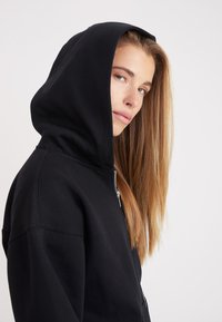 Joven mujer con cabello largo y rubio que lleva puesta una sudadera negra con cremallera y capucha, mirando de lado contra un fondo liso.