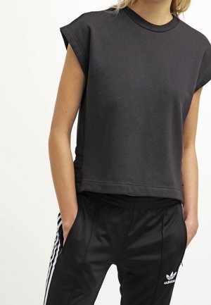 Kvinde iført en ærmeløs, mørkegrå cropped top og sorte Adidas træningsbukser med hvide sidetstriber, hænderne i lommerne.