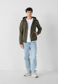 Giacca verde oliva con zip e cappuccio, arricchita da dettagli testurizzati. Abbinata a una maglietta bianca e jeans azzurri chiari, con vestibilità comoda.