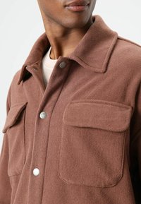 Bruine fleece overhemdjas met een kraag, twee voorzakken en zilveren drukknoopsluitingen, met een zachte textuur en een ontspannen pasvorm.