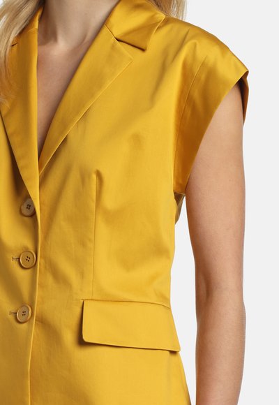 Ujjatlan mustársárga blézer három gombbal és egy zsebfedővel, amelyet világos bőrű és szőke hajú személy visel.