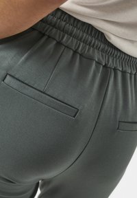 Pantalone tamnozelene boje s elastičnim pojasom, s dva stražnja džepa i glatkom, mat teksturom. Fokus na prilagođeni dizajn pantalona.