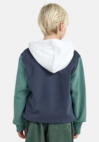 Hoodie met een marineblauwe achterkant, groene mouwen en een witte capuchon. Zacht materiaal met een ontspannen pasvorm en geribbelde manchetten.