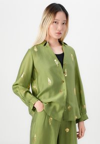 Junge Frau mit langen blonden Haaren, die ein lockeres olivgrünes Hemd und eine Hose mit goldenen Muschelmuster trägt, steht vor einem weißen Hintergrund.