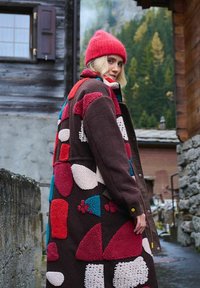 Osoba v patterned vlnenom kabáte a červenej pletenej čiapke vonku blízko rustikálnych drevených budov a zalesnených hôr.