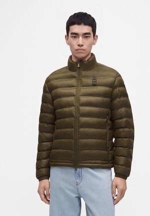 Homme portant une veste matelassée vert olive avec une fermeture éclair et un jean bleu clair, debout devant un fond blanc uni.