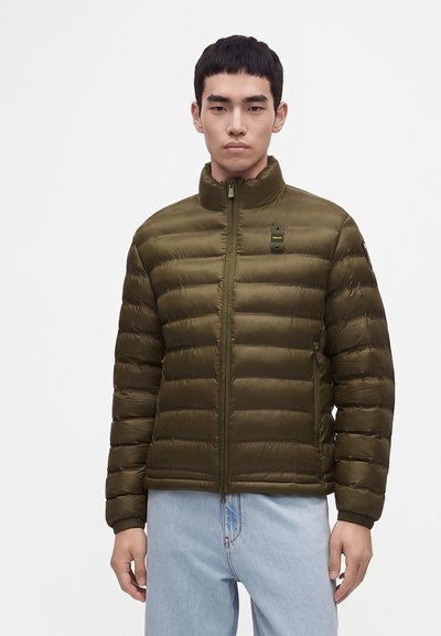 Homme portant une veste matelassée vert olive avec une fermeture éclair et un jean bleu clair, debout devant un fond blanc uni.