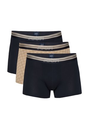 Passender Mann mit kurzen dunklen Haaren steht barfuß und trägt schwarze Boxershorts mit grauem Bund vor schlichtem hellem Hintergrund.