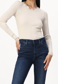 Beige ribbat långärmad topp med spetsdetalj runt halsringningen, matchad med mörkblå slim-fit jeans med femfickorsdesign.