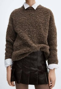 Maglione marrone a texture con scollo rotondo, indossato sopra una camicia bianca a righe con colletto. Abbinato a una gonna plissettata nera in pelle.