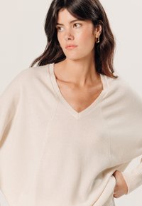 Lichtbeige V-halstrui met een losse pasvorm en geribbelde halslijn. De stof lijkt zacht, met subtiele naden en een relaxte silhouet.