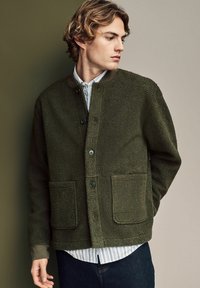 Olive green textured fleece jacket featuring a round collar, two front pockets, and button fastenings, worn over a striped shirt.