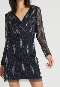 Námořnické šaty s dlouhými průsvitnými rukávy, zdobené složitými stříbrnými korálky ve vlnkových vzorech a výstřihem do V. Krátká délka a těsný střih.