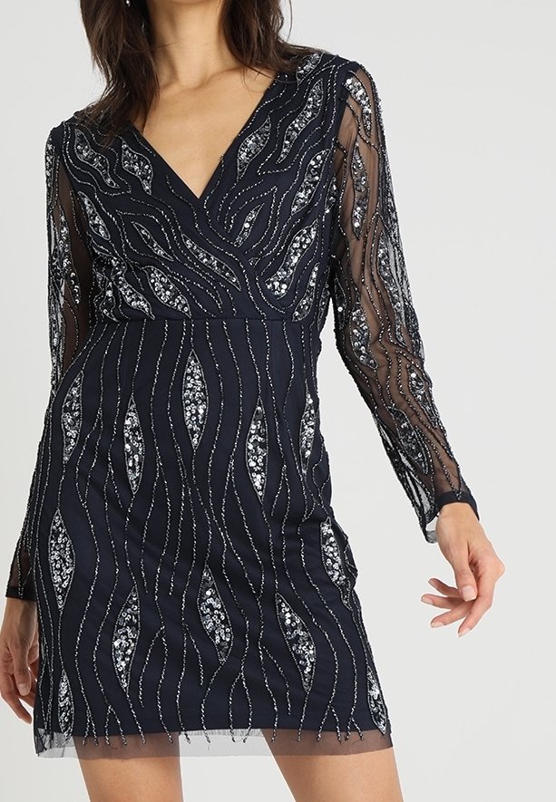 Námořnické šaty s dlouhými průsvitnými rukávy, zdobené složitými stříbrnými korálky ve vlnkových vzorech a výstřihem do V. Krátká délka a těsný střih.