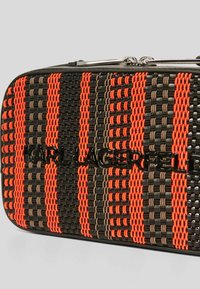 Geweven handtas met zwarte, oranje en bruine strepen, met een gladde textuur en een glanzend zwart logo op de voorkant. Ritssluiting aan de bovenkant.