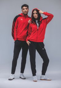 Des sweat-shirts à capuche zippés rouges avec des accents noirs, associés à des pantalons noirs. Logo présenté sur les sweat-shirts. Modèles portant des baskets blanches. Fond simple.