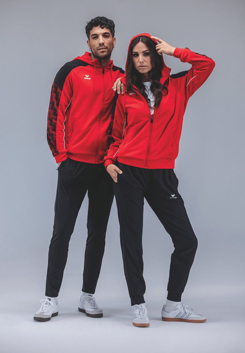 Rode zip-up hoodies met zwarte accenten, gecombineerd met zwarte broeken. Kenmerkend logo op de hoodies. Modellen dragen witte sneakers. Eenvoudige achtergrond.