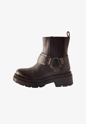 Braune Leder-Ankleboots mit elastischen Seiteneinsätzen, einer dicken schwarzen Sohle und einem dekorativen Riemen mit einem Metallring. Flaches Absatzdesign.