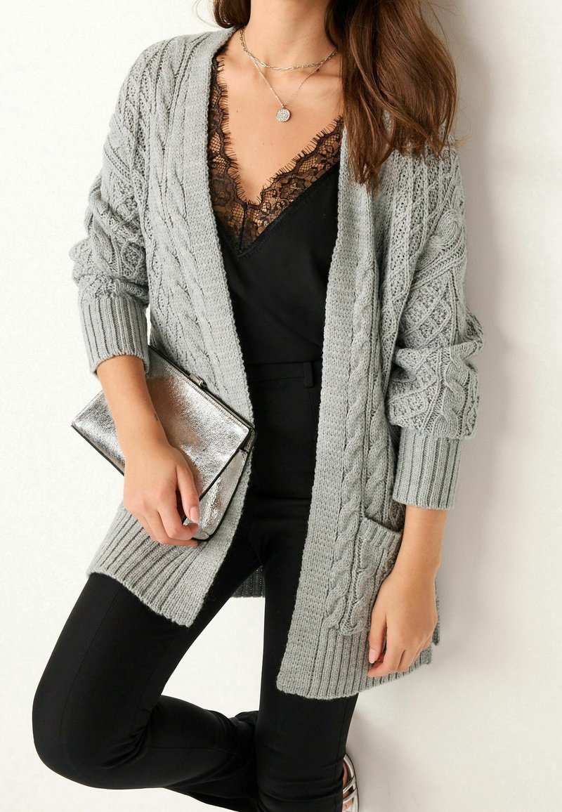 Femme portant un cardigan gris en maille torsadée sur un top noir orné de dentelle, tenant une pochette argentée, avec un pantalon noir et un collier pendentif.